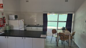 A compact kitchen with white cabinets, a black countertop, a microwave, and a refrigerator. There is a dining area with a table and chairs, and a window with curtains. - Alice Motor Inn (East Side)