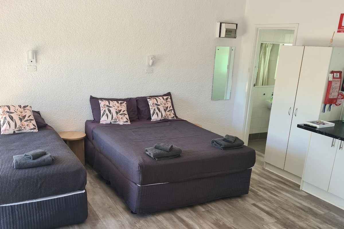 A modern bedroom with two beds, a wooden floor, and a kitchenette in the background.