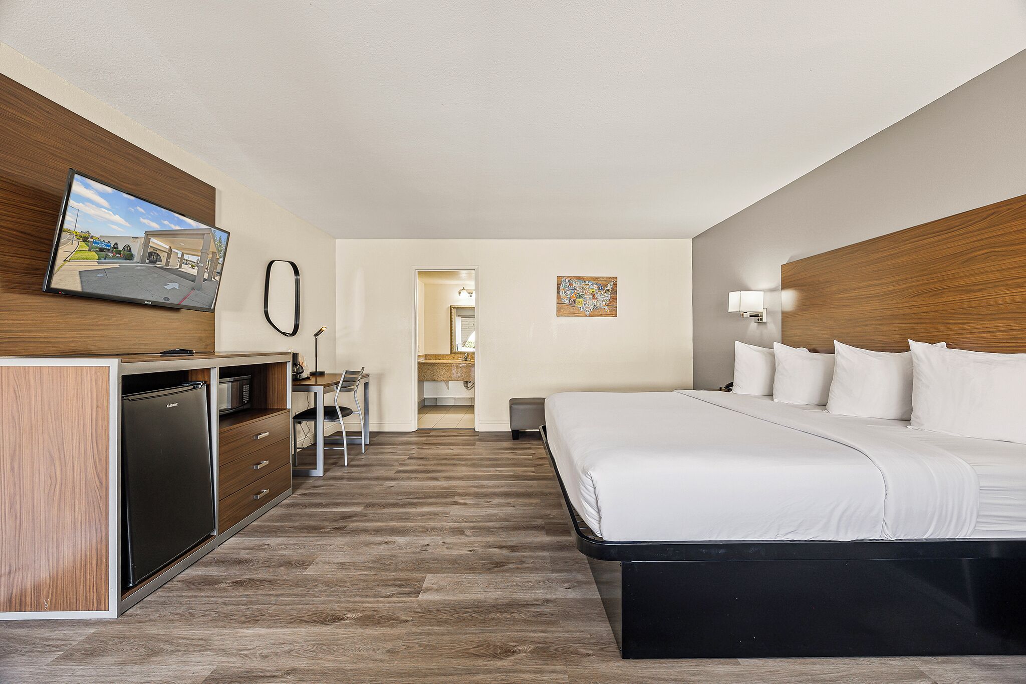 Habitació d'hotel amb un llit, una escriptori amb un ordinador, un refrigerador, una televisió i un bany visible per una porta oberta.
