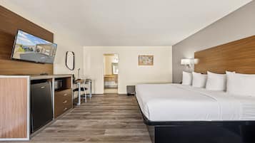 A hotel room with a bed, a desk with a computer, a refrigerator, a television, and a bathroom visible through an open door.