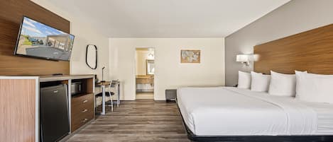 Habitació d'hotel amb un llit, una escriptori amb un ordinador, un refrigerador, una televisió i un bany visible per una porta oberta.