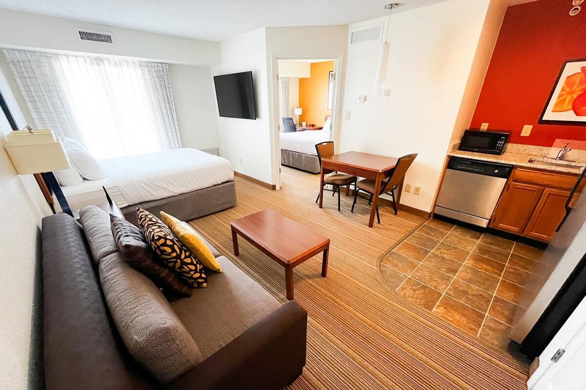 Une chambre d’hôtel comprenant un lit, un canapé, une table à manger, un coin cuisine et un téléviseur fixé au mur.