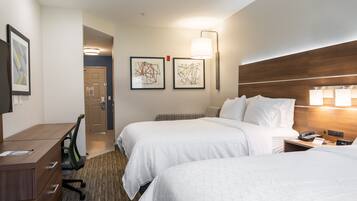 A hotel room with two beds, a desk, a chair, and a lamp.