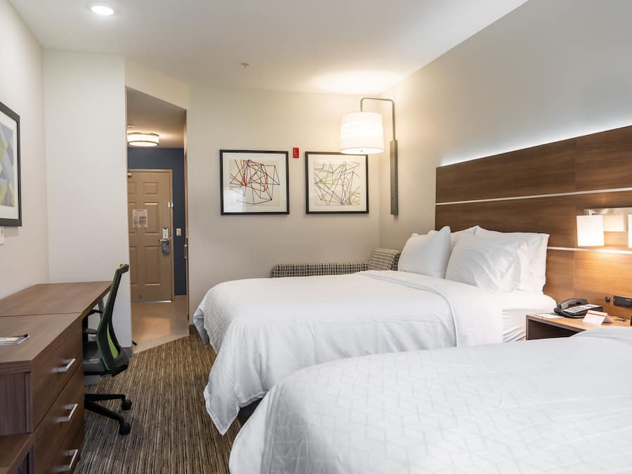 A hotel room with two beds, a desk, a chair, and a lamp.