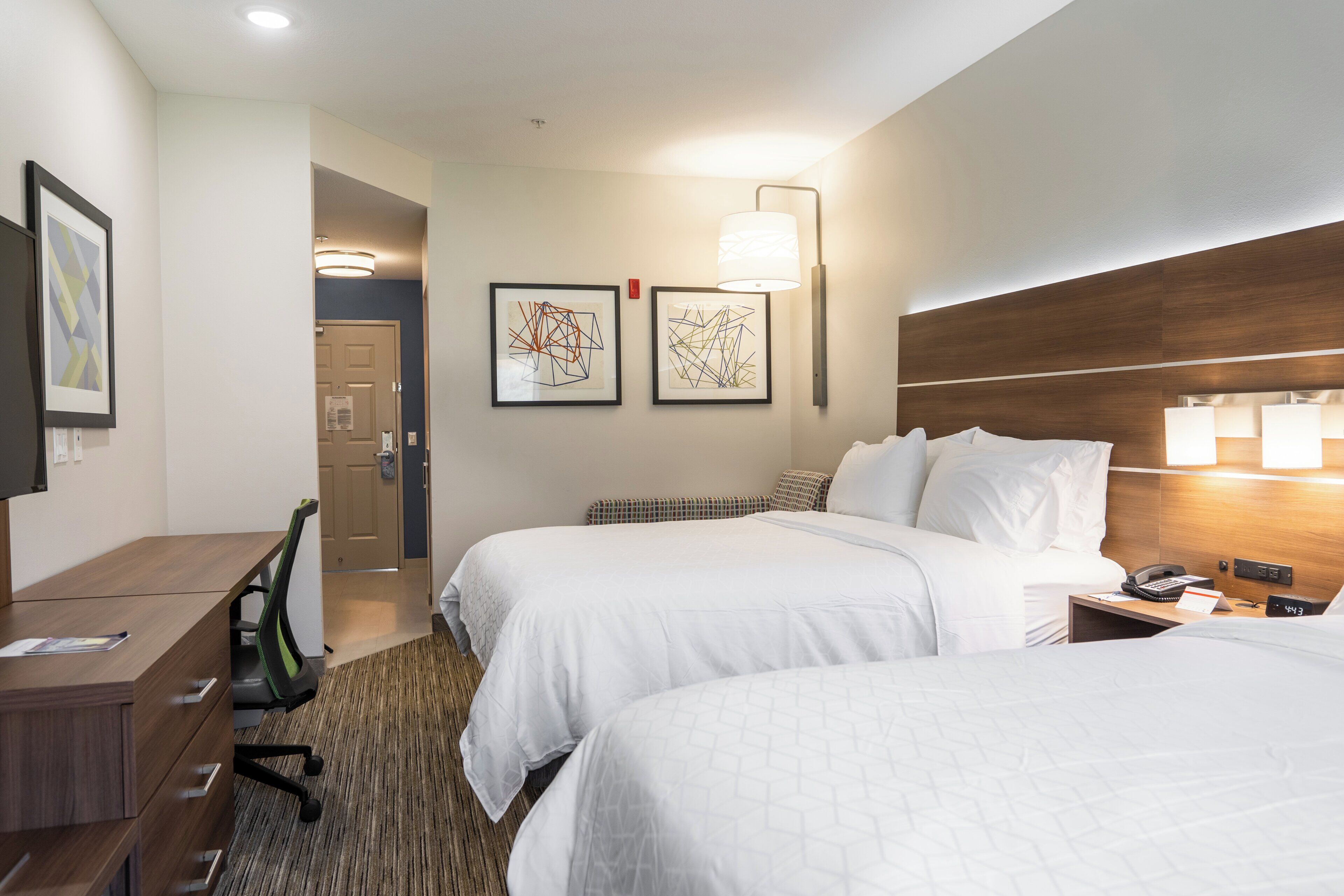 A hotel room with two beds, a desk, a chair, and two framed abstract artworks on the wall.