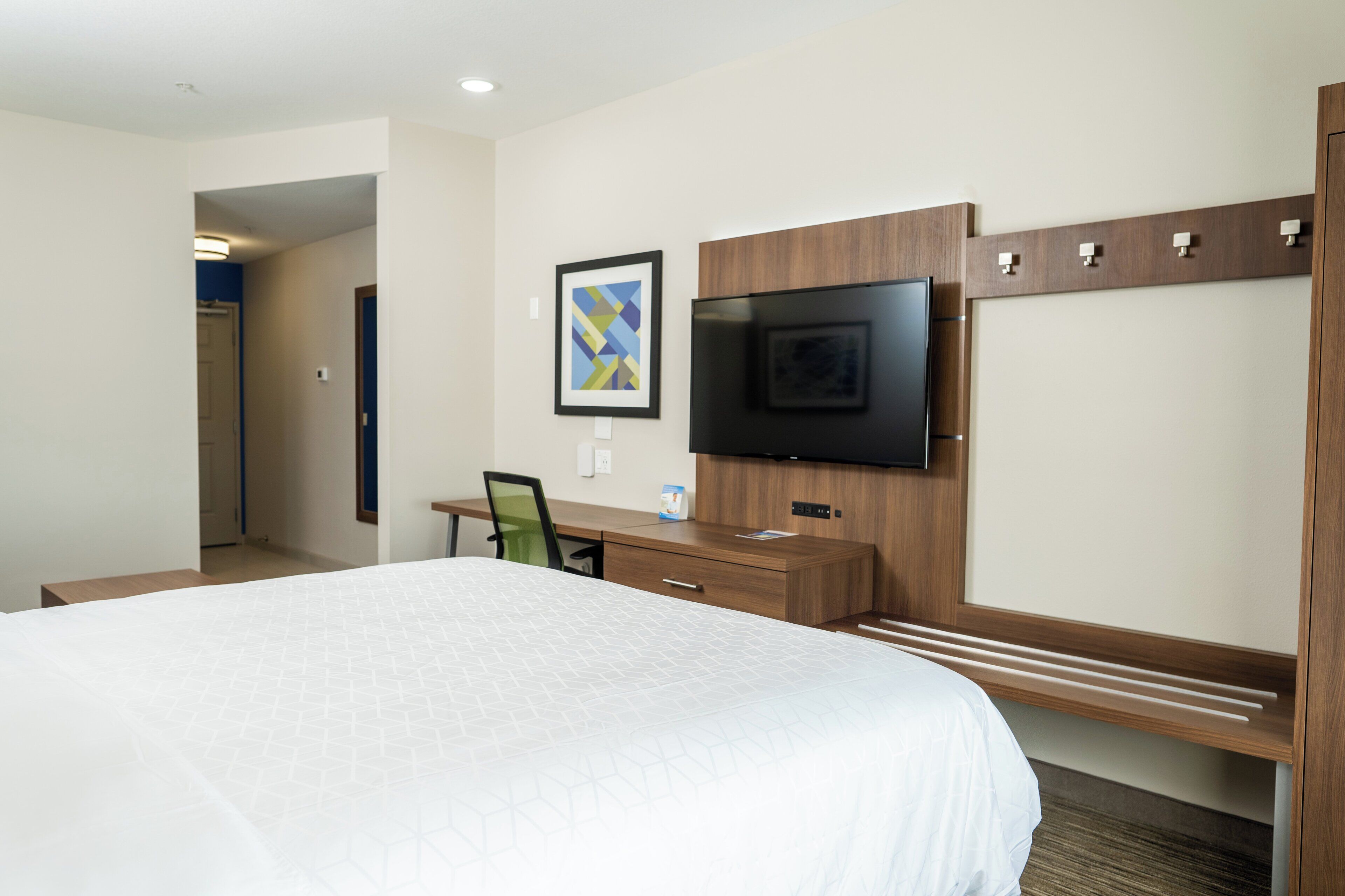 Habitación de hotel con una cama grande, un escritorio, una silla, un televisor de pantalla plana montado en la pared y un cuadro enmarcado sobre el televisor.