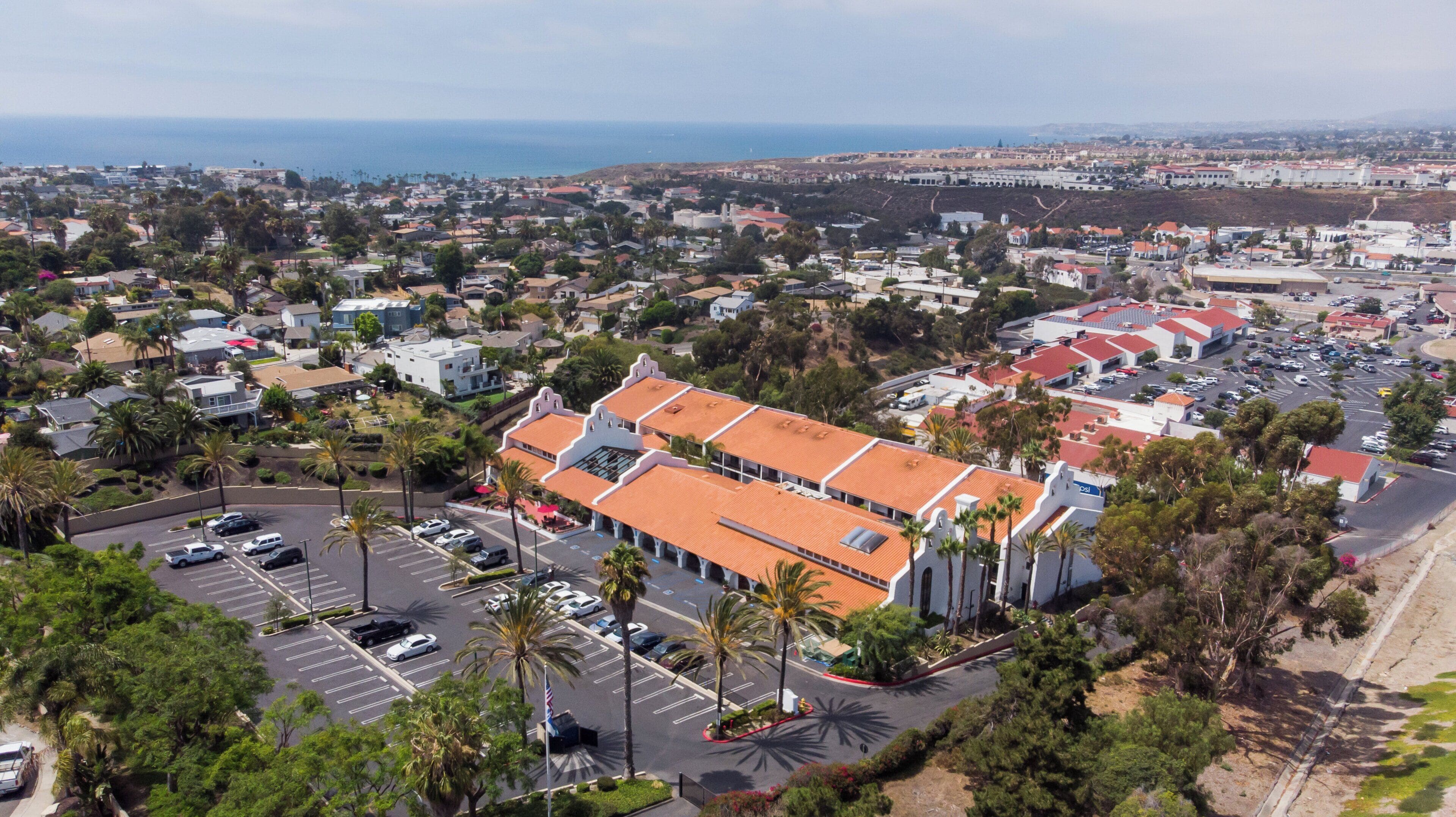 An aerial view of a coastal town with a prominent building featuring a red-tiled roof, surrounded by parking spaces and palm trees.