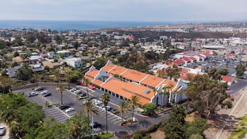 An aerial view of a coastal town with a prominent building featuring a red-tiled roof, surrounded by parking spaces and palm trees.