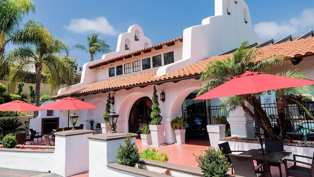 A building with a white facade, red-tiled roof, and arched windows. There are outdoor tables and red umbrellas, and palm trees in the background. - Holiday Inn Express San Clemente N – Beach Area by IHG (San Clemente)