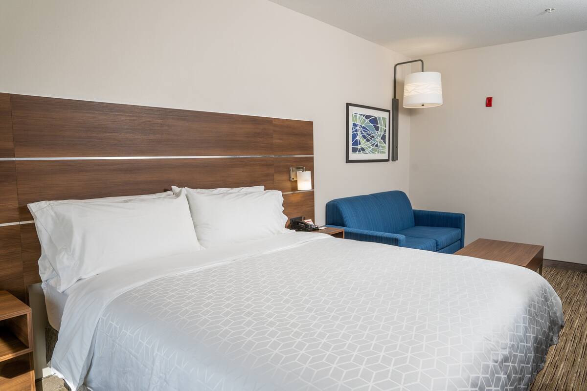 A hotel room with a large bed, a blue armchair, a nightstand, and a wall-mounted lamp.