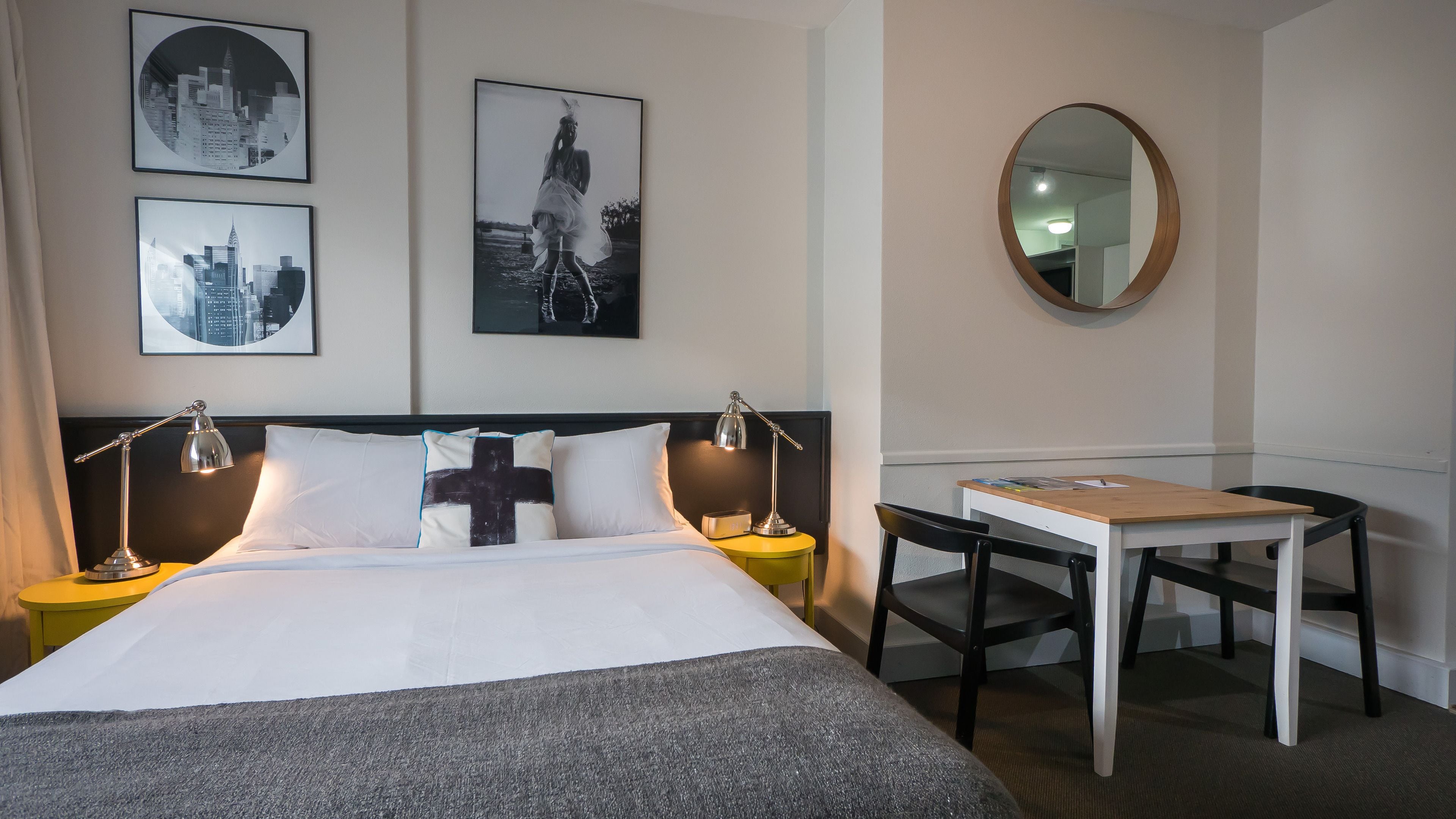 A hotel room with a bed, two bedside tables with lamps, a desk with a chair, and two framed pictures on the wall.