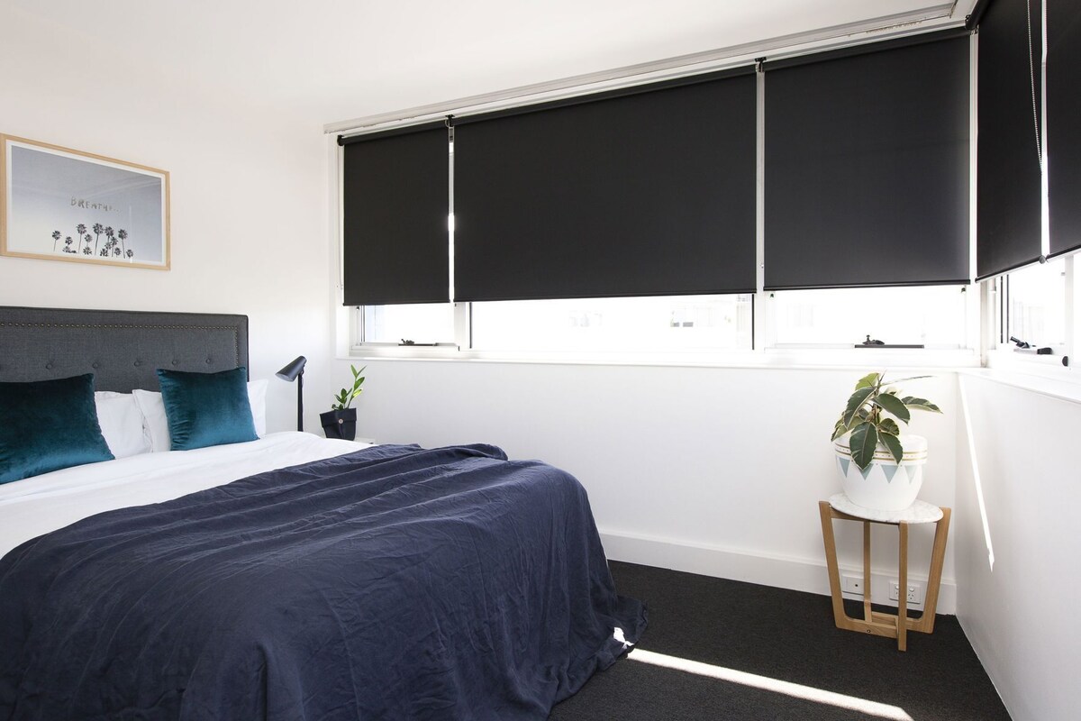 Een slaapkamer met een bed, een nachtkastje, een potplant en een raam met jaloezieën.