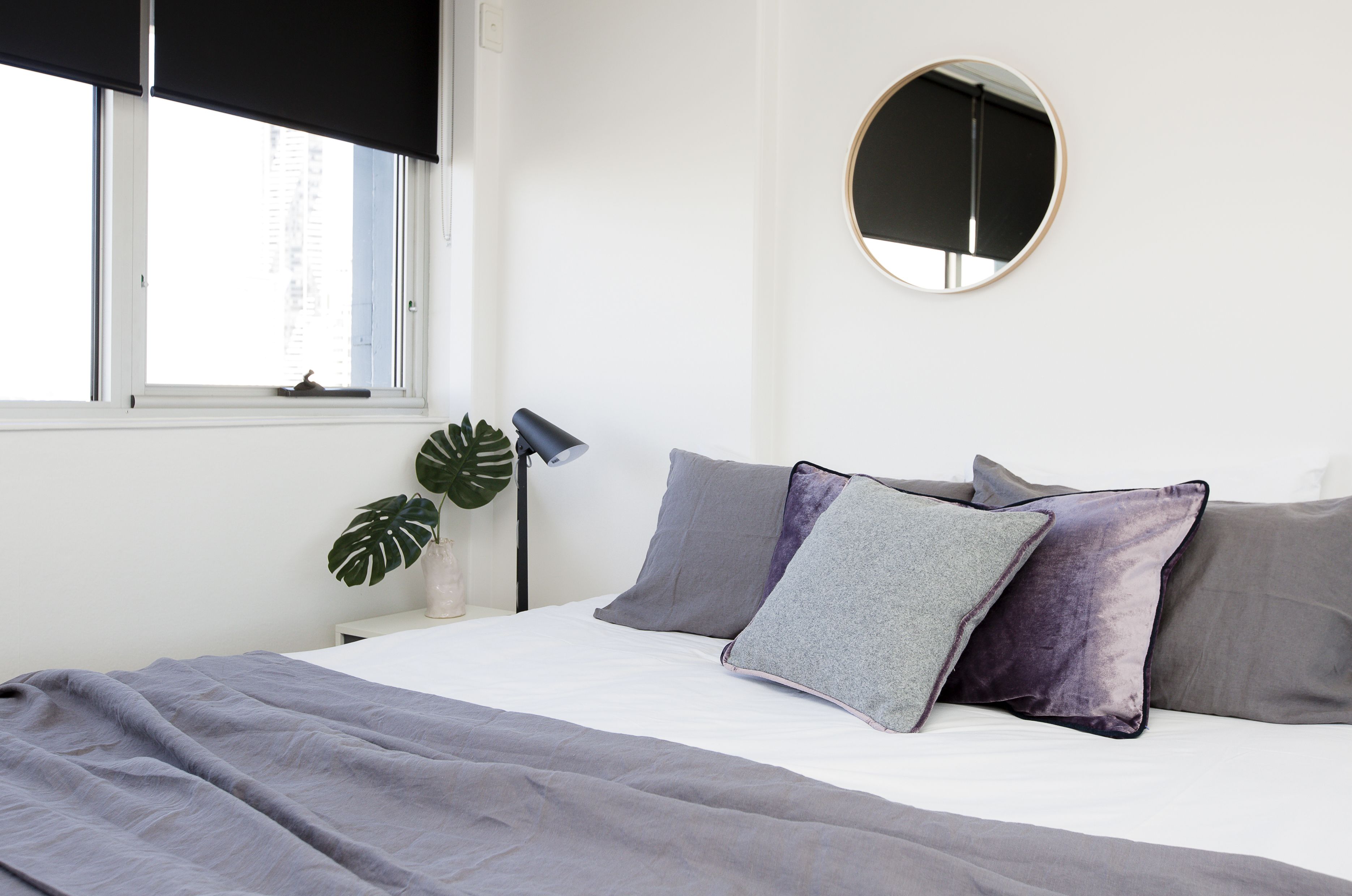 Un lit bien fait, avec une literie grise, un miroir rond accroché au mur et une plante placée près de la fenêtre.
