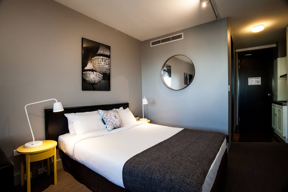 Un lit bien fait, avec du linge de lit blanc et un plaid gris, un miroir rond accroché au mur, une lampe moderne posée sur une table de chevet jaune, et une œuvre d’art encadrée au-dessus du lit.