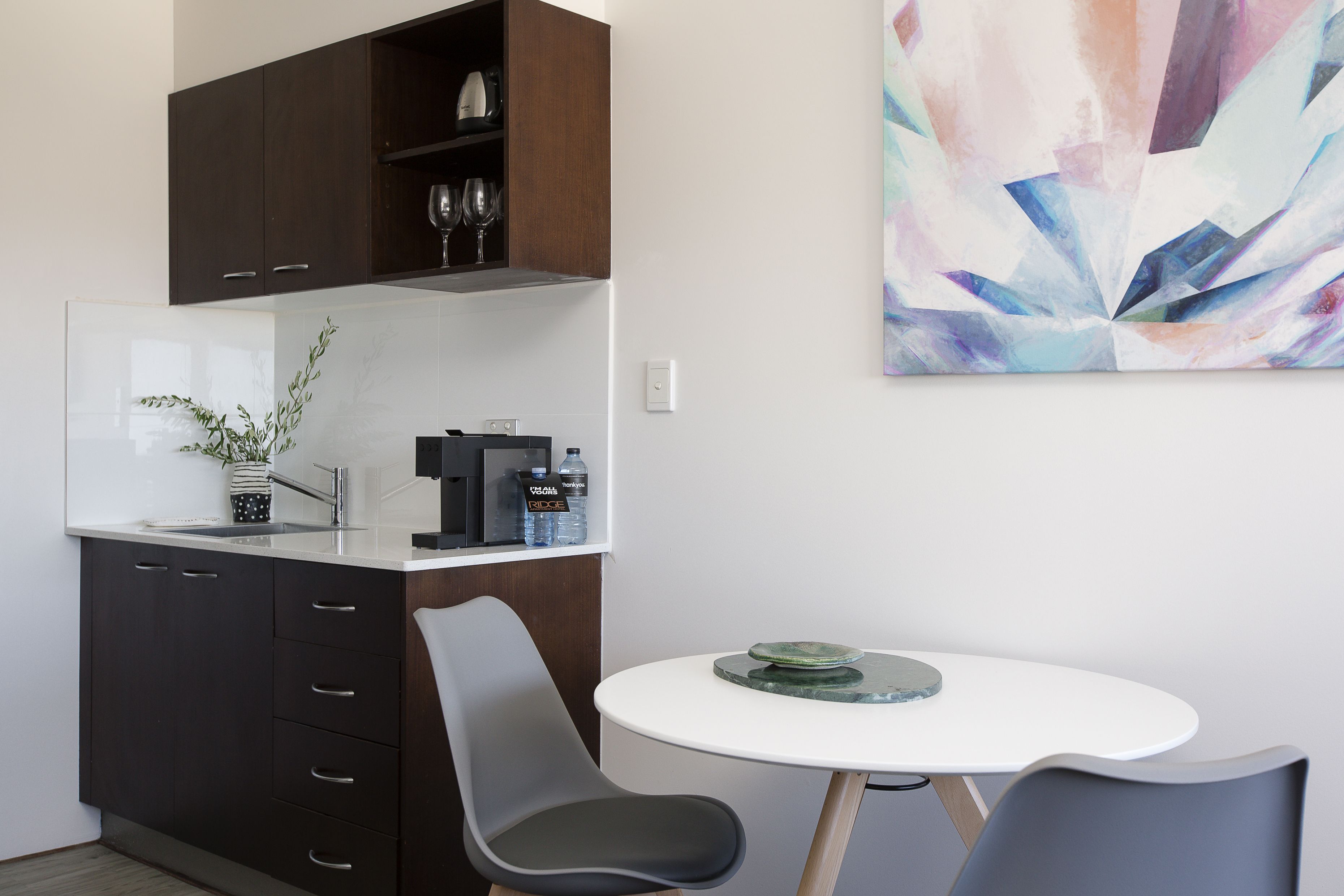 A modern kitchen with dark cabinets, a white countertop, and a coffee machine. There is a dining area with a round table and two chairs.