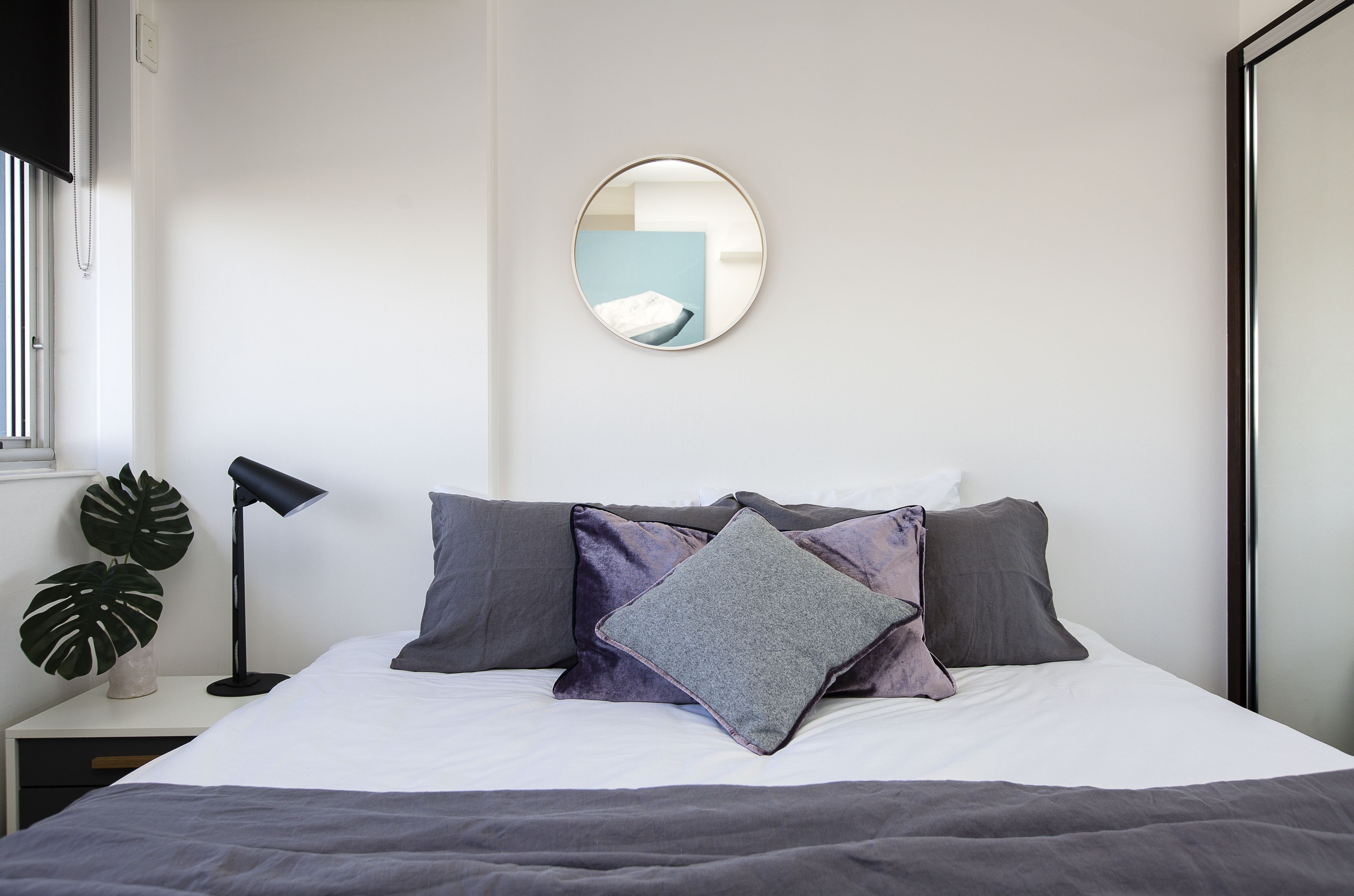 A neatly made bed with grey and purple bedding, a round mirror on the wall, and a black floor lamp beside the bed.