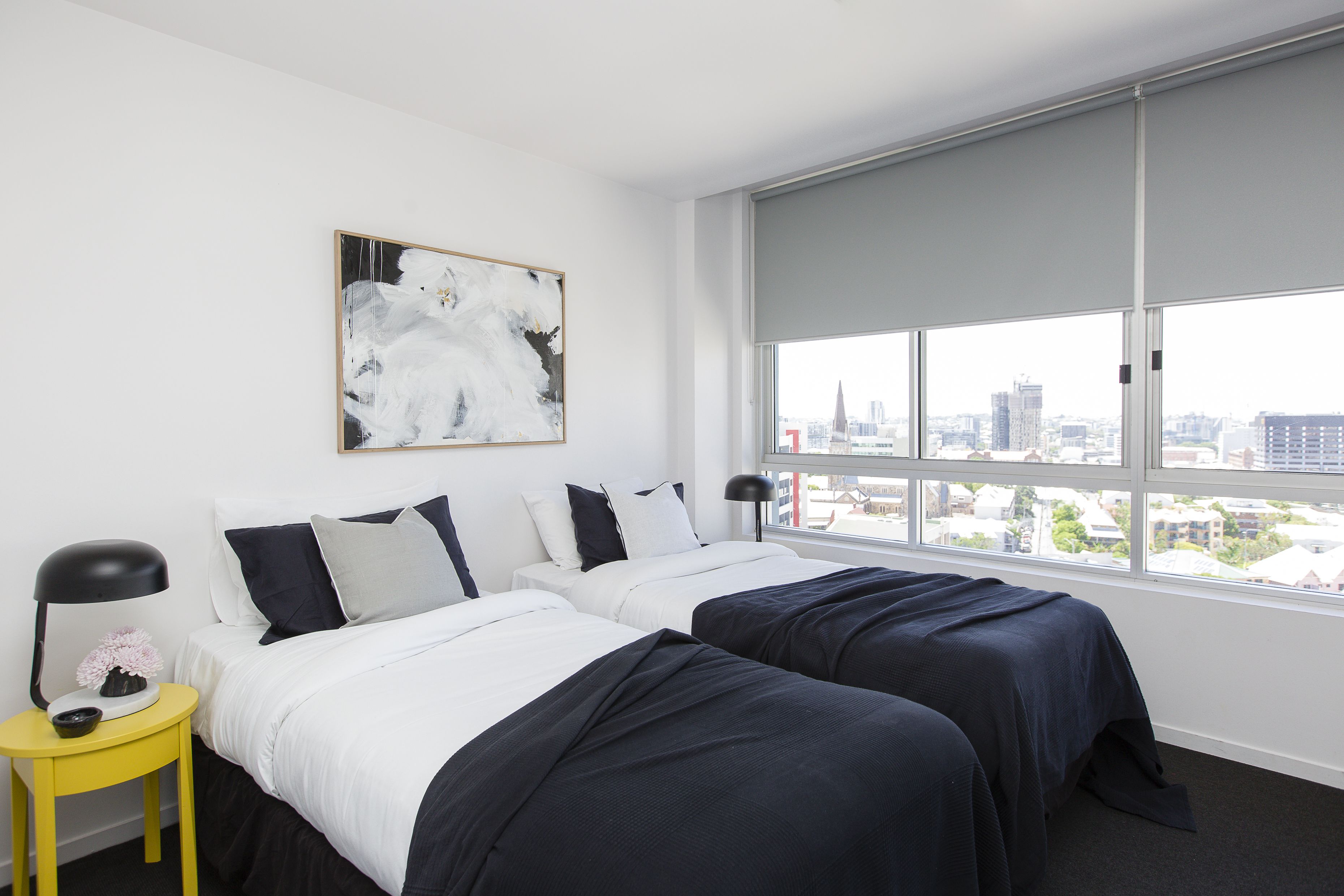 A hotel room with two beds, a large window with a city view, a yellow side table, and a modern abstract painting.