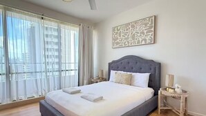 Une chambre à coucher avec un grand lit, un ventilateur de plafond, une fenêtre avec des rideaux légers et une œuvre d’art accrochée au mur.