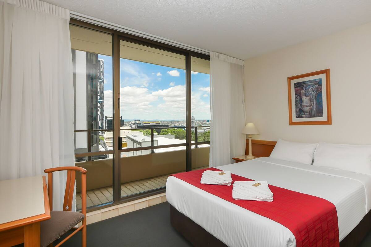 A hotel room with a large window, a bed with white and red bedding, a desk, a chair, and a nightstand with a lamp.