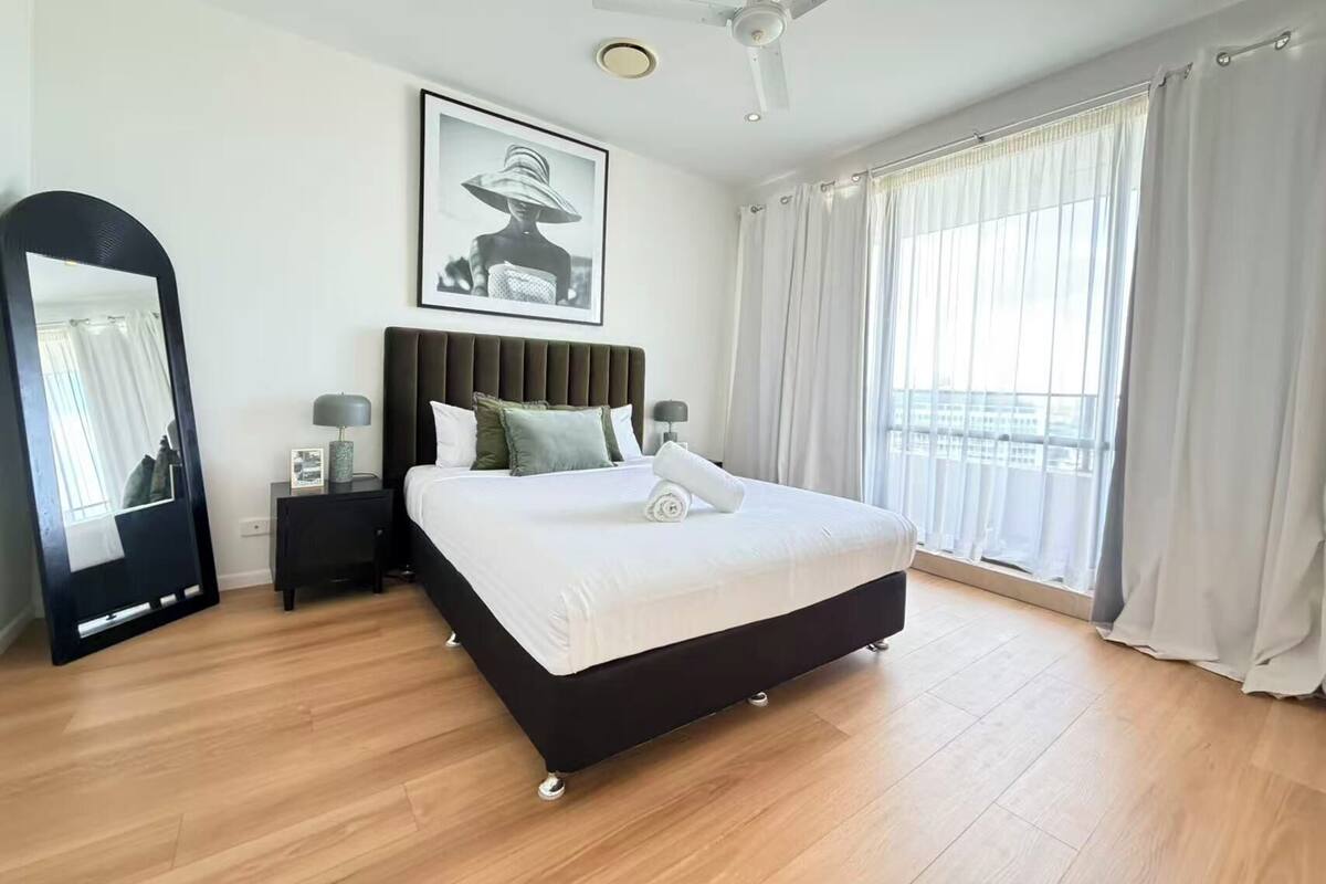 A modern bedroom with a bed, a ceiling fan, a large window with curtains, a wall-mounted mirror, and a framed picture.