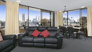 Une chambre d’hôtel moderne avec un canapé en cuir noir, des coussins rouges, un coin repas avec des chaises et de grandes fenêtres donnant sur la ville.