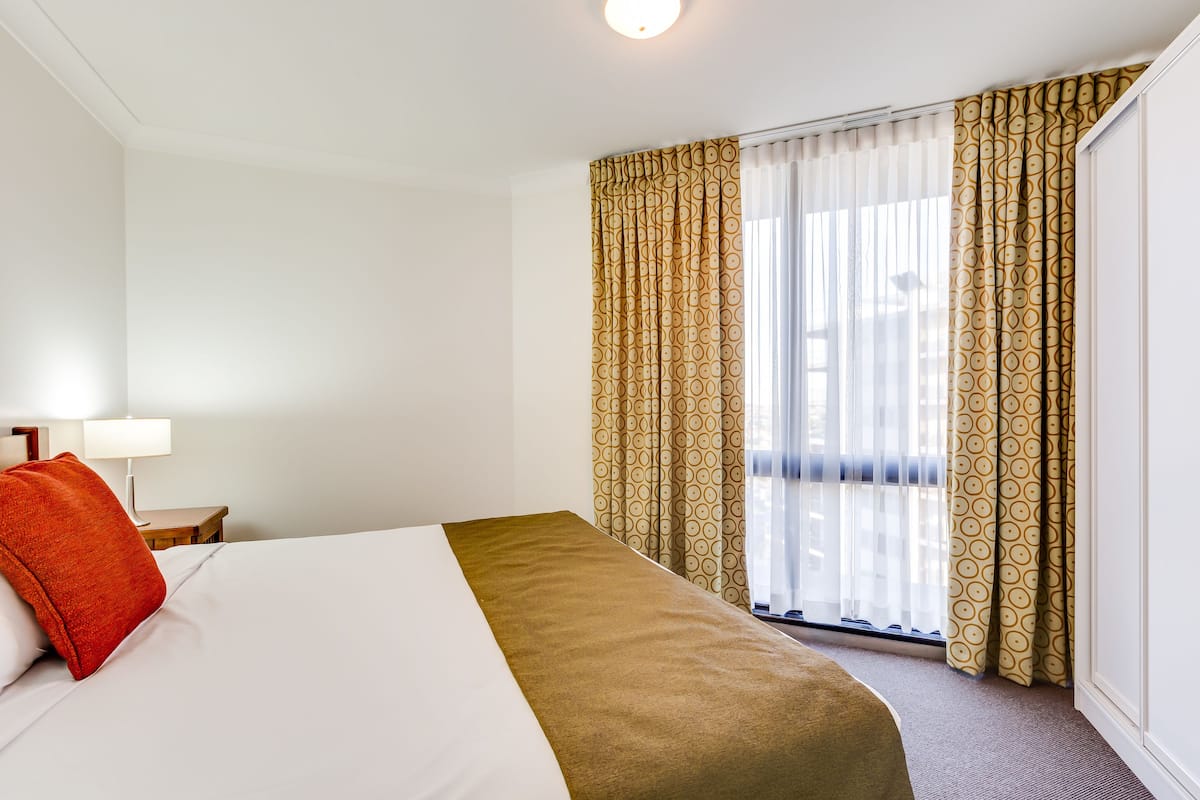 Habitación de hotel con cama, mesita de noche, lámpara y ventana con cortinas.