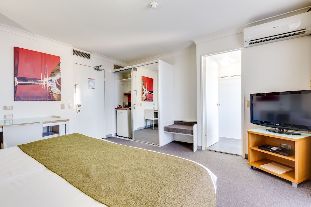 Una habitación de hotel con cama, televisión, escritorio y baño visible a través de una puerta abierta.