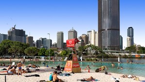 Una playa con gente tomando el sol y nadando, una torre de salvavidas y un horizonte urbano al fondo.