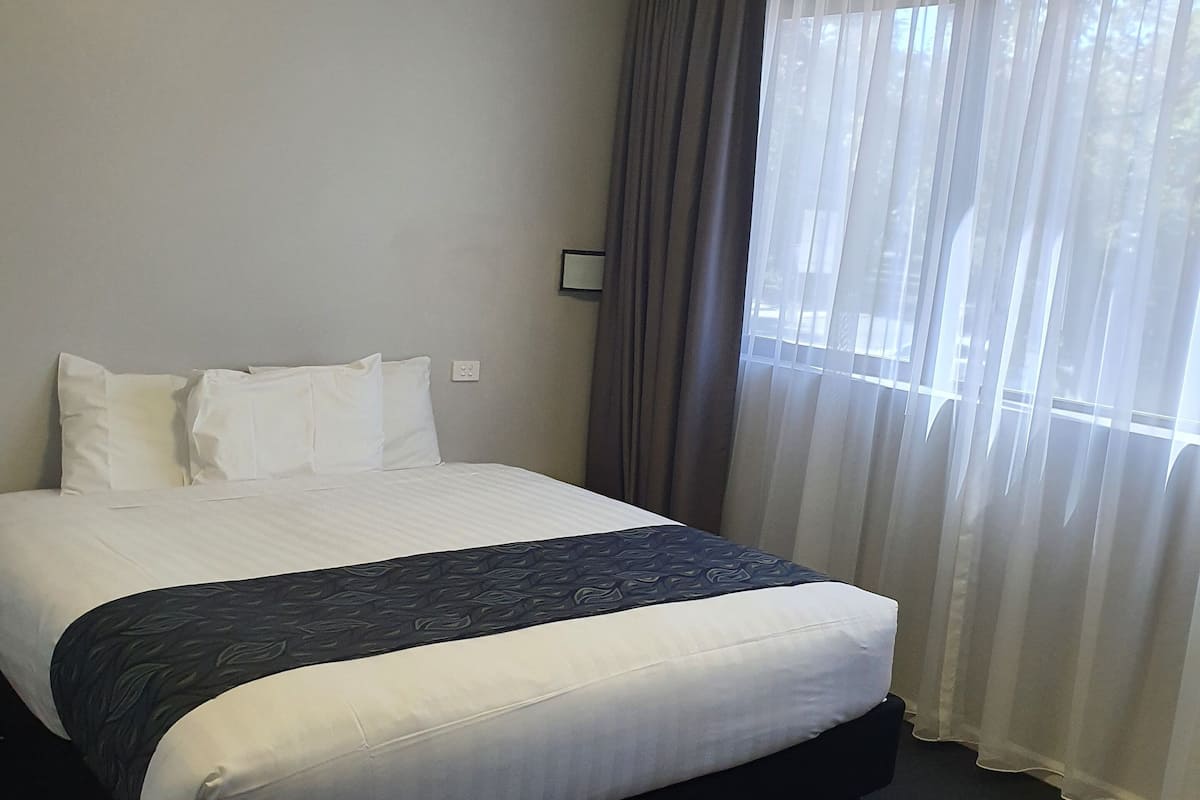 A hotel room with a large bed, a dark carpet, a window with sheer curtains, and a plain wall.