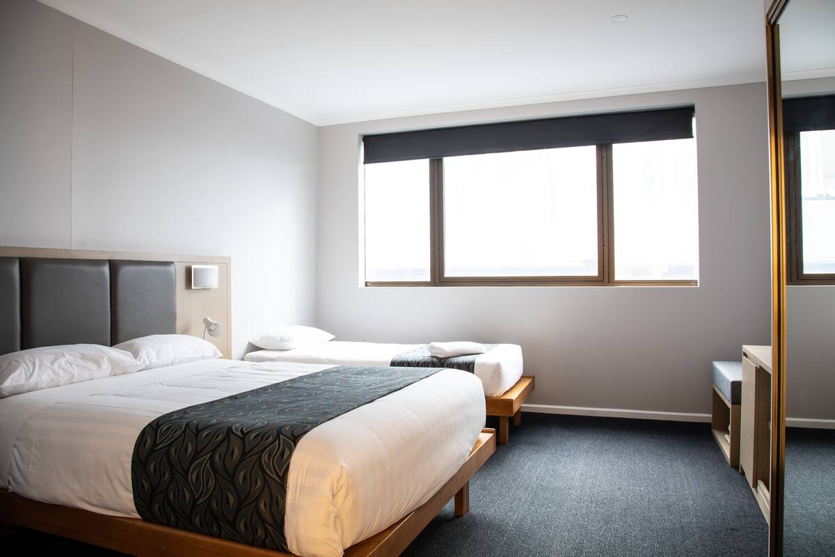 A hotel room with a large bed, a smaller bed, a desk, and a window with blinds.