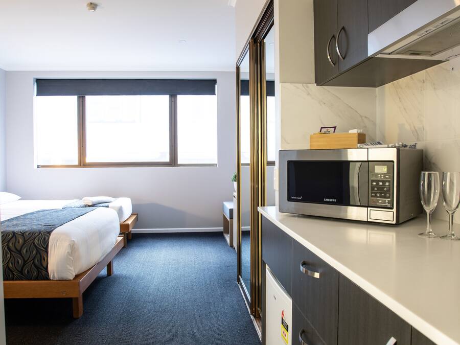 Quarto de hotel com cama, micro-ondas, garrafa de vinho e uma pequena mesa.
