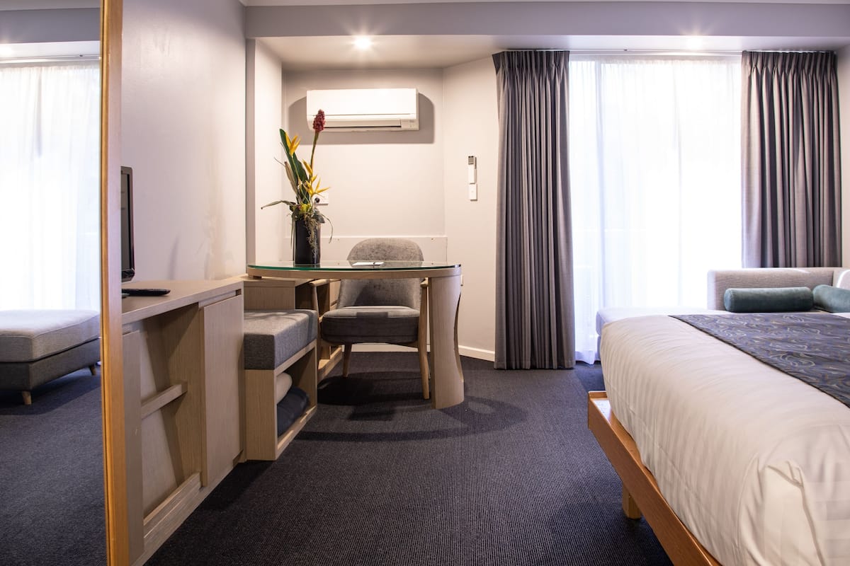 A hotel room with a bed, a desk with a chair, a small table, and a potted plant.