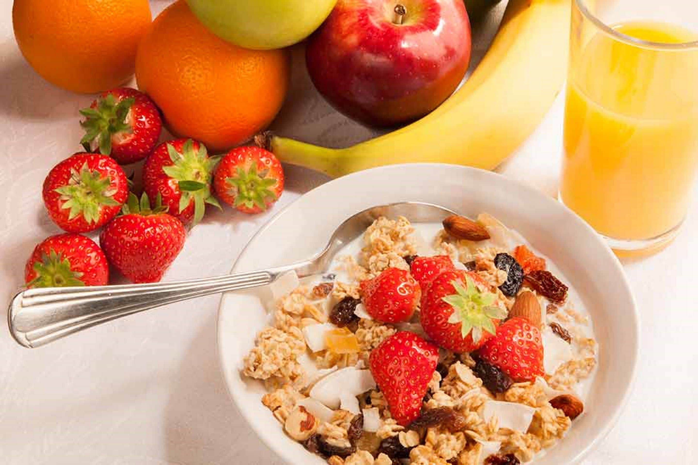 A bowl of cereal with strawberries and almonds, surrounded by fresh fruits and a glass of orange juice.