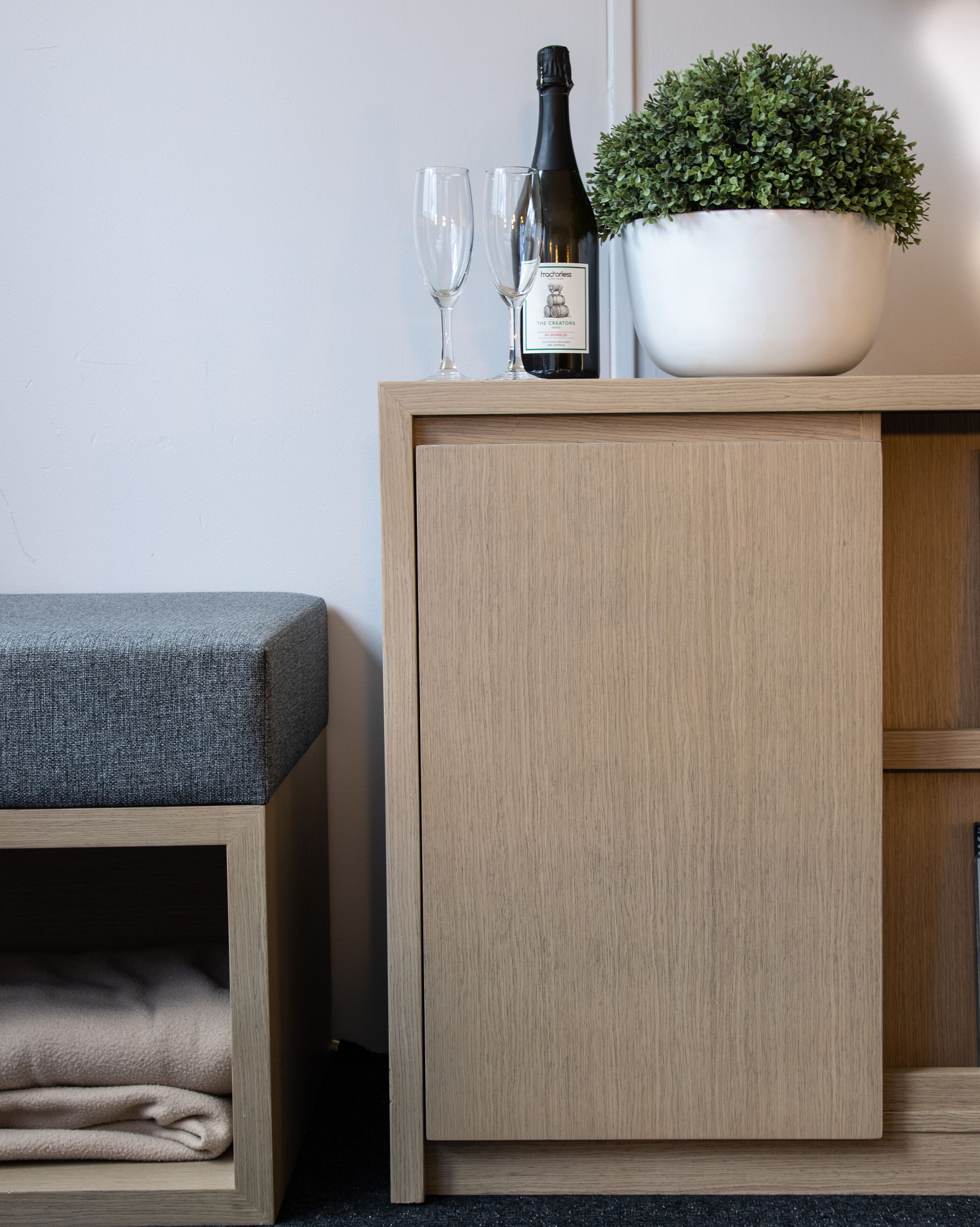 A wooden cabinet with a bottle of wine and two glasses on top, a potted plant, and a grey cushioned bench.