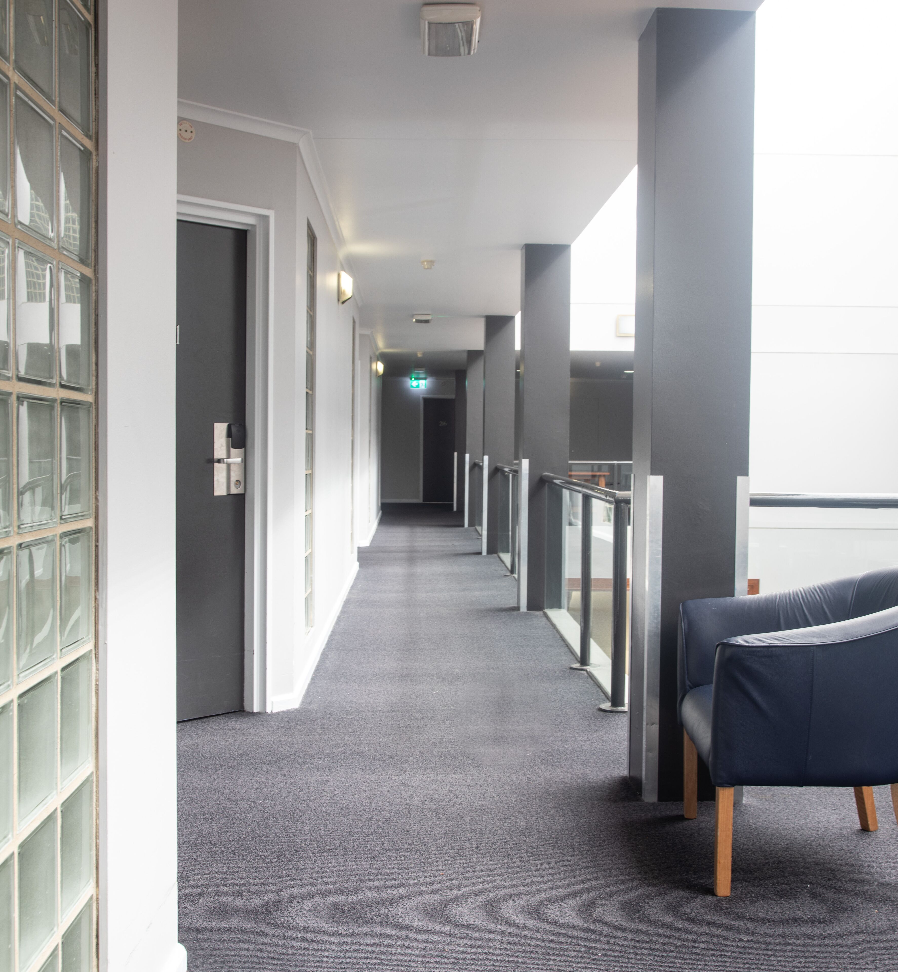 Un couloir avec un tapis gris, une porte à gauche et une chaise bleue à droite.