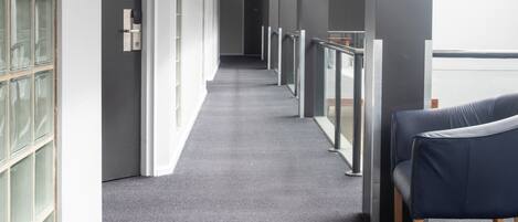 Un couloir avec un tapis gris, une porte à gauche et une chaise bleue à droite.
