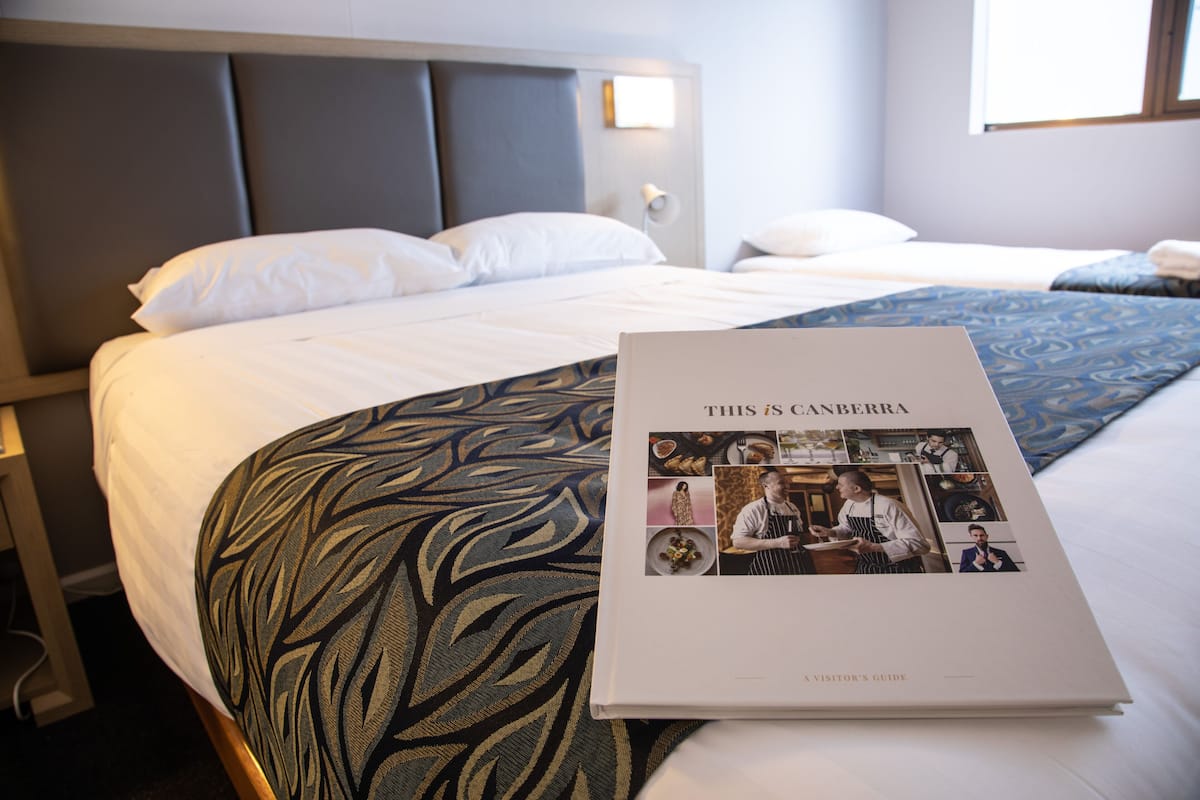 A hotel room with a neatly made bed, a book titled "THIS IS CANBERRA," and a patterned throw blanket.
