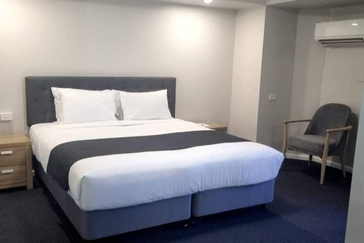 A neatly arranged hotel room with a large bed, a bedside table, a chair, and an air conditioning unit.