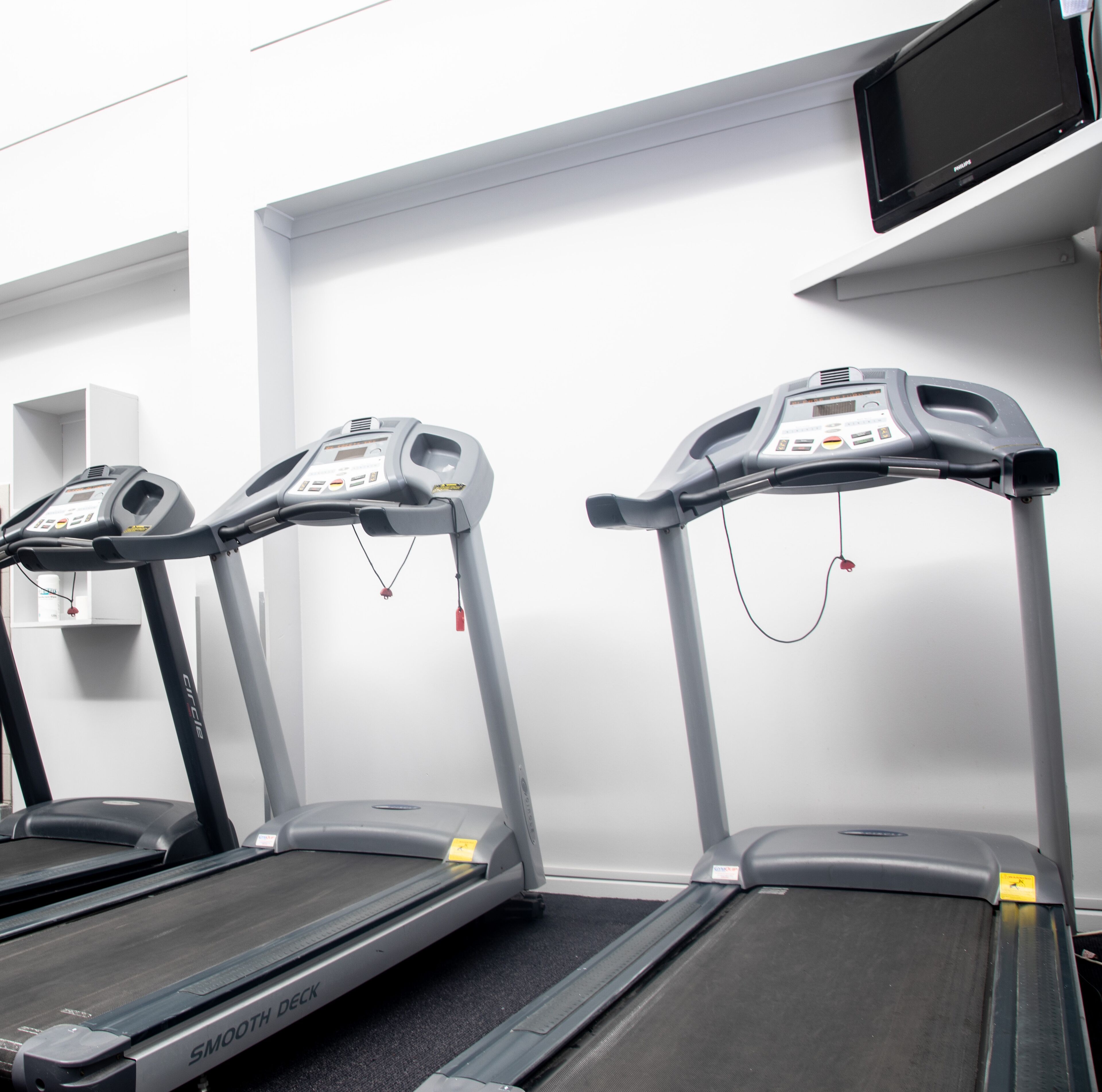Three treadmills in a gym with a white wall and a television mounted above.