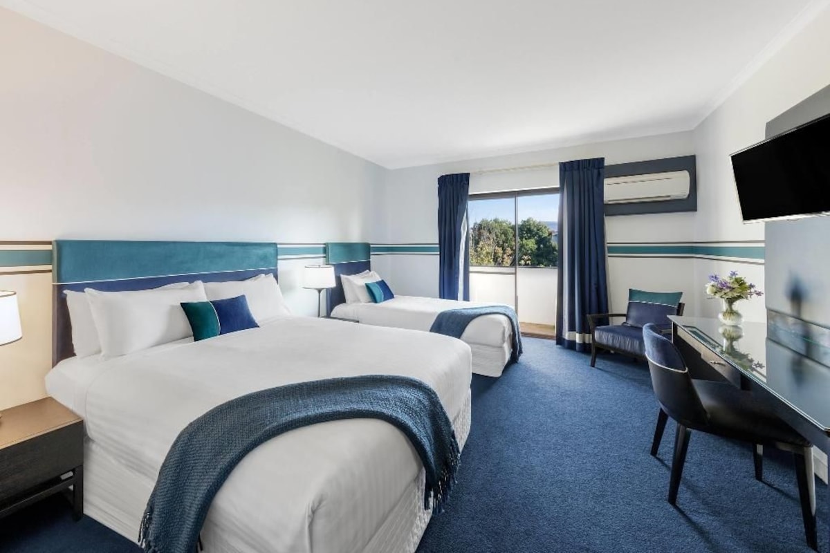 A hotel room with two beds, a desk, a chair, a TV, and a window with curtains.