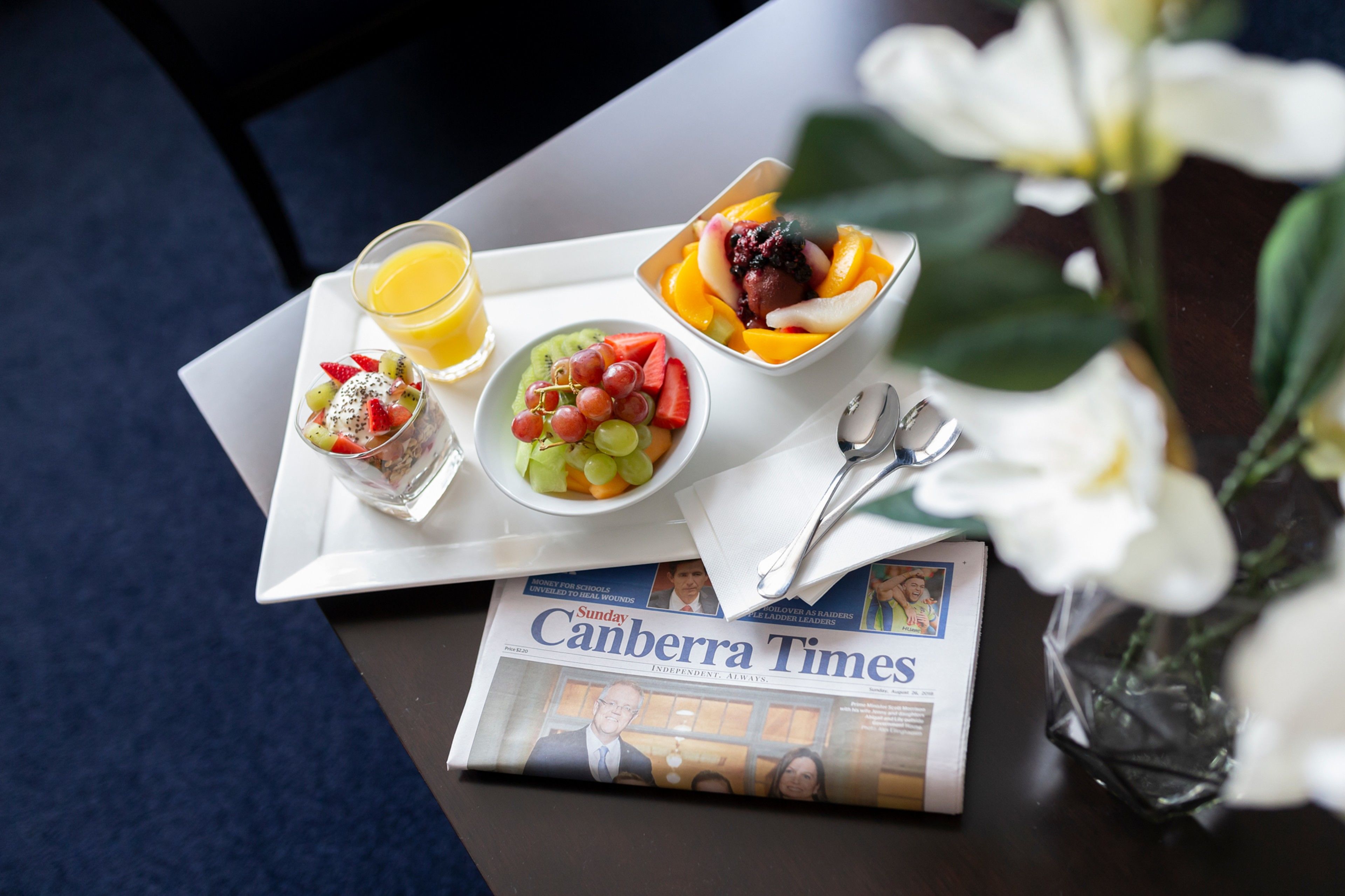 Ein Frühstücksbrett mit verschiedenen Früchten, einem Orangensaftglas, einer Zeitung und einem Löffel auf einem Tisch.