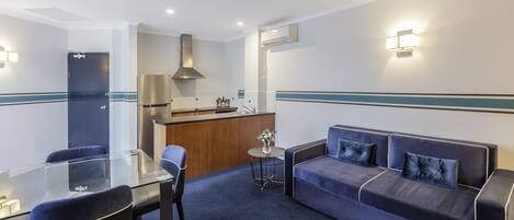 A modern hotel room with a glass dining table, a sofa, and a small kitchen area.