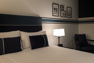 Un cuarto de hotel con una cama, una mesita de noche con lámpara, una silla y cuadros en la pared.