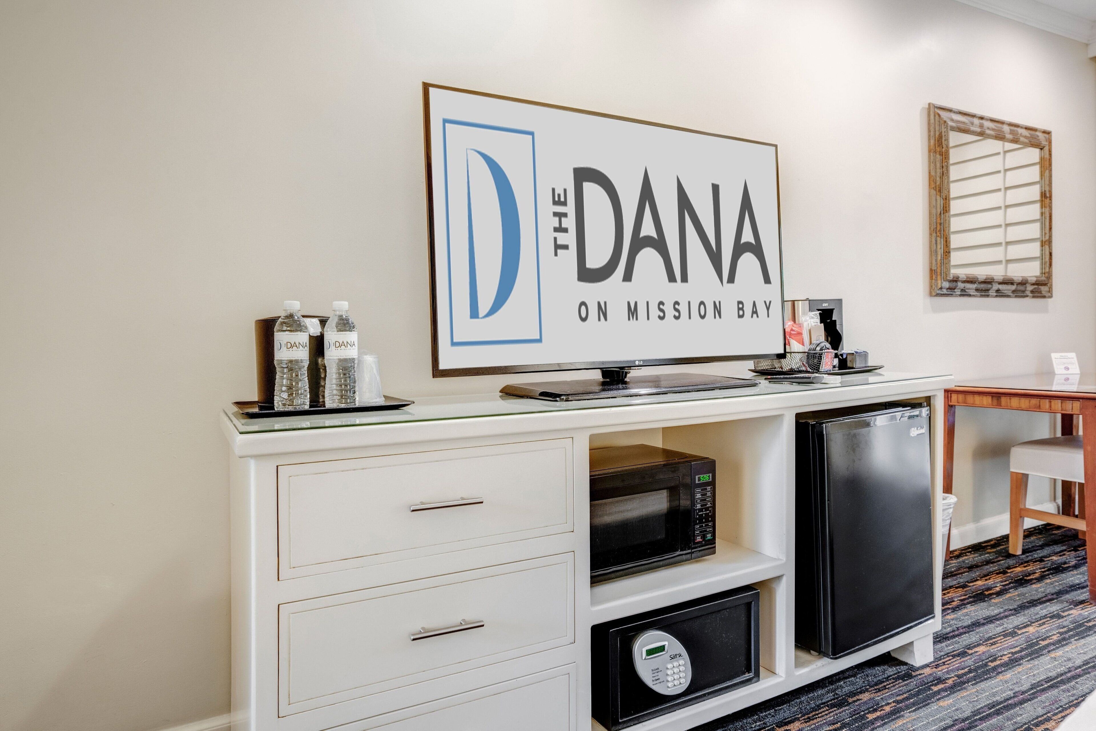 A kitchenette with a microwave, refrigerator, and a coffee maker on a white cabinet. A framed sign above reads "The Dana on Mission Bay."