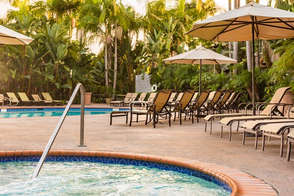 Zona de piscina con sillones de playa y sombrillas, rodeada de palmeras.