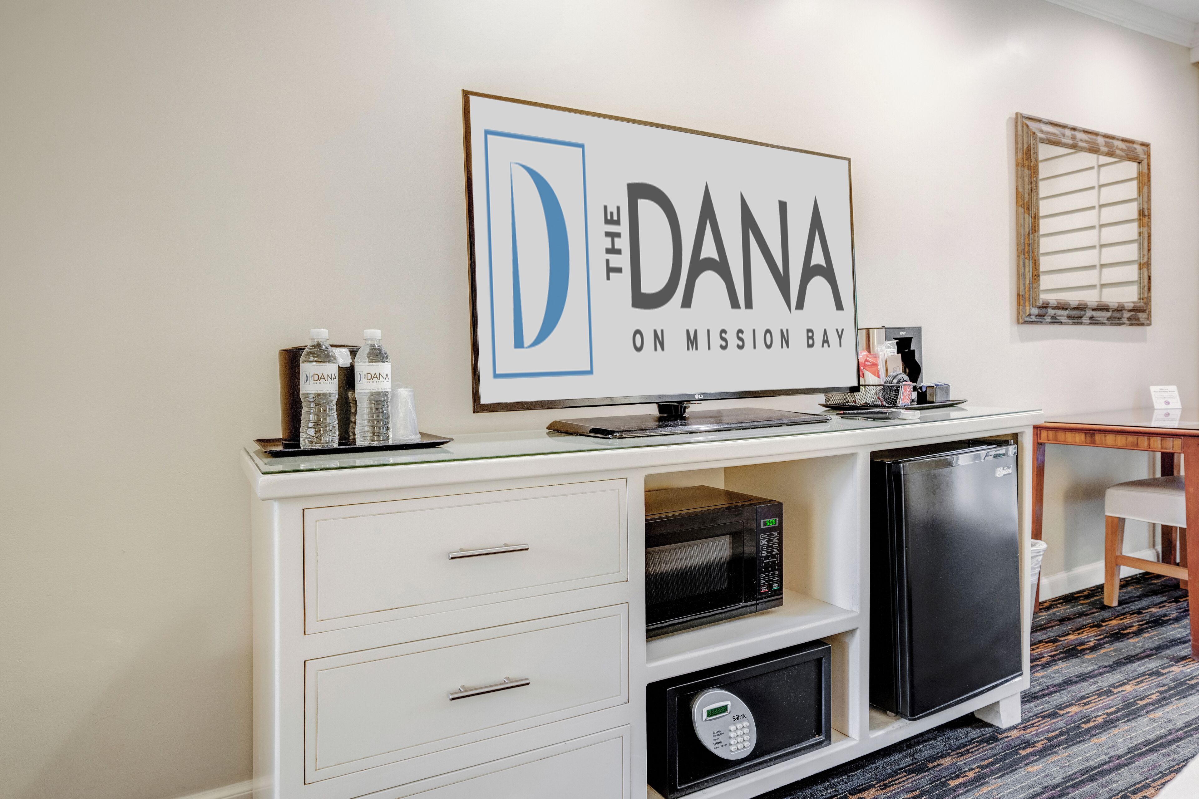 A kitchenette with a microwave, refrigerator, and a coffee maker on the counter. There is a framed sign above the counter that reads "The Dana on Mission Bay."