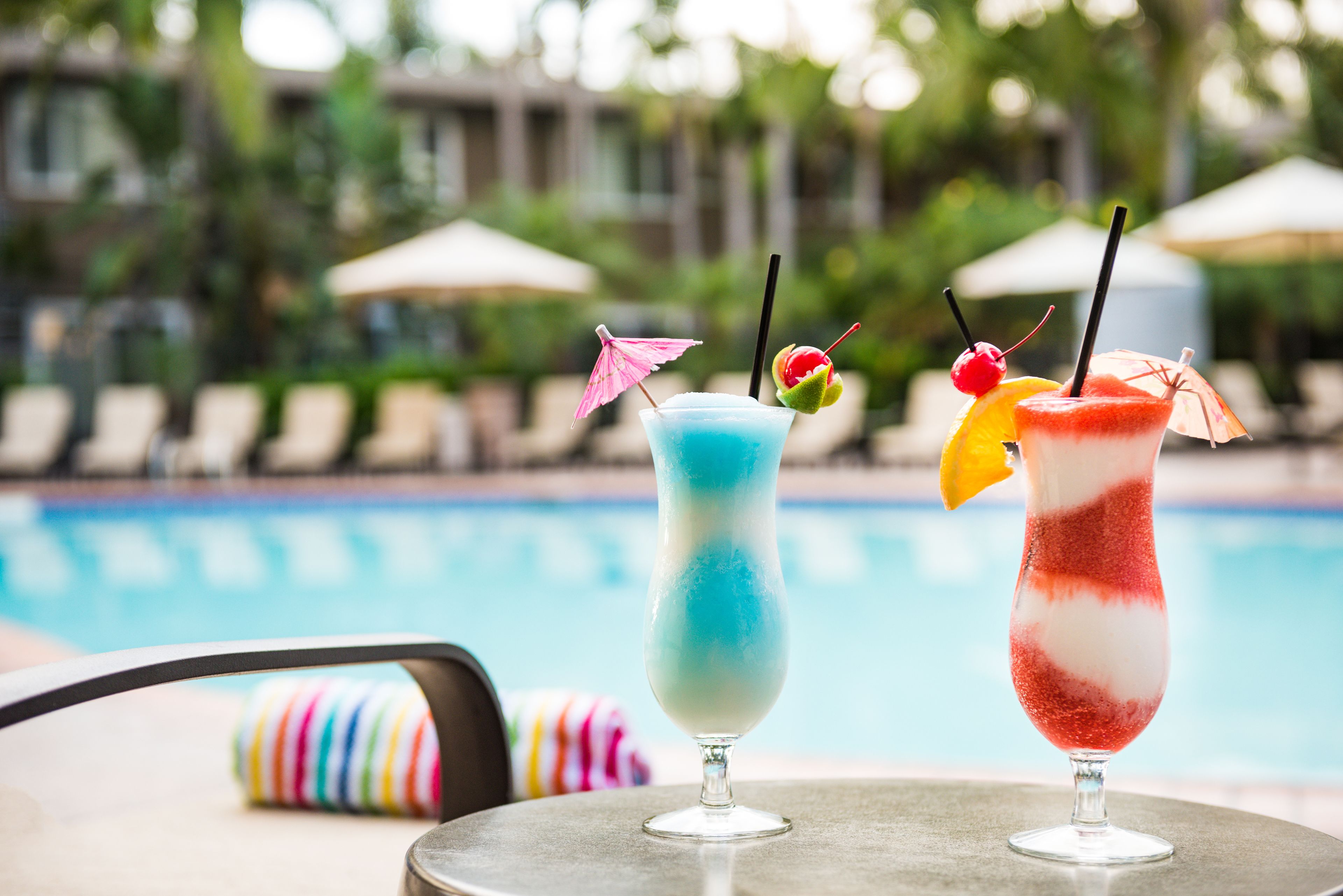 Two cocktails on a table by a poolside with lounge chairs and umbrellas in the background.