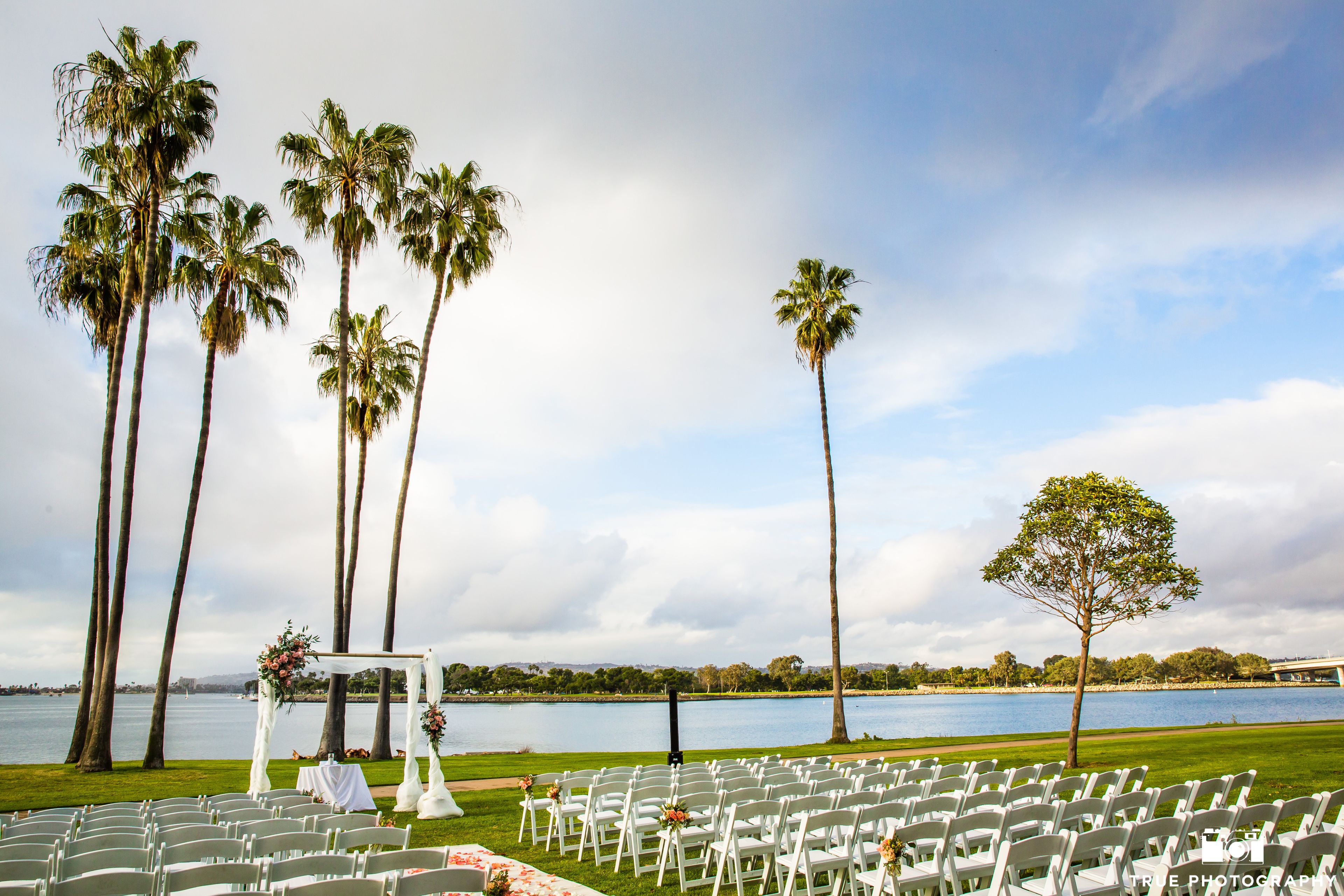 A wedding ceremony setup with white chairs and a white arch, palm trees, and a lake view.