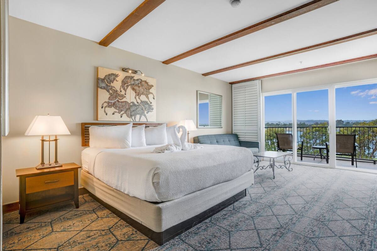 A hotel room with a large bed, a balcony with chairs, and a scenic view.