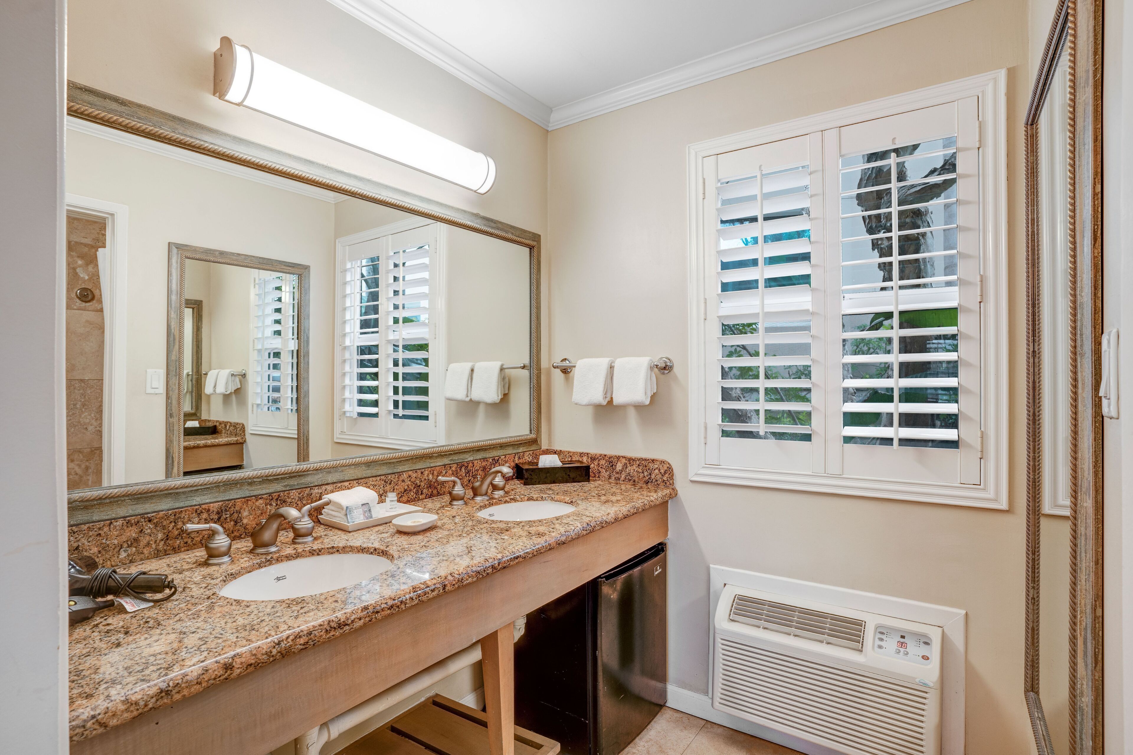 A bathroom with a granite countertop, two sinks, a large mirror, and a window with blinds.